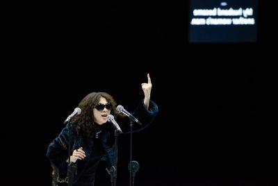 An actress, an Asian woman with long red-brown hair, wears sunglasses and a fur coat and speaks into three microphones in stands, her right hand pointing towards the ceiling.